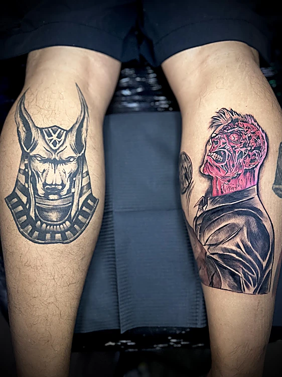 Dos tatuajes en las pantorrillas: Anubis en negro y gris y el villano Dos Caras con la mitad del rostro en rojo.