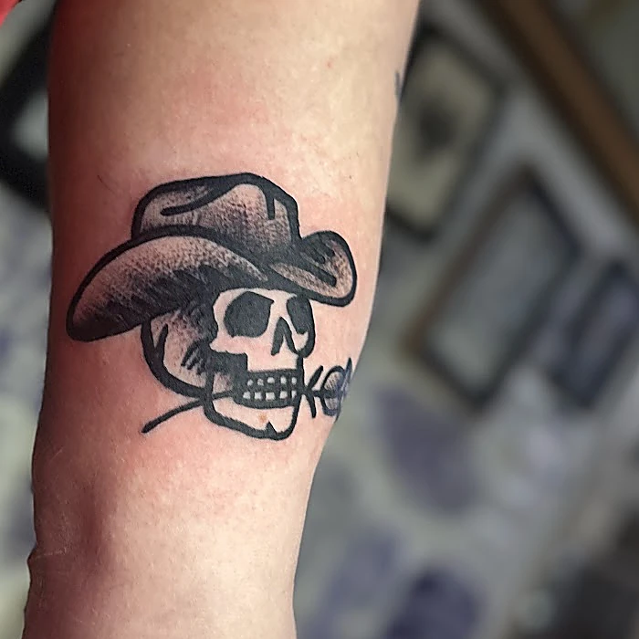 Tatuaje tradicional en el brazo: calavera con sombrero vaquero y una flor en la boca, en negro sólido.