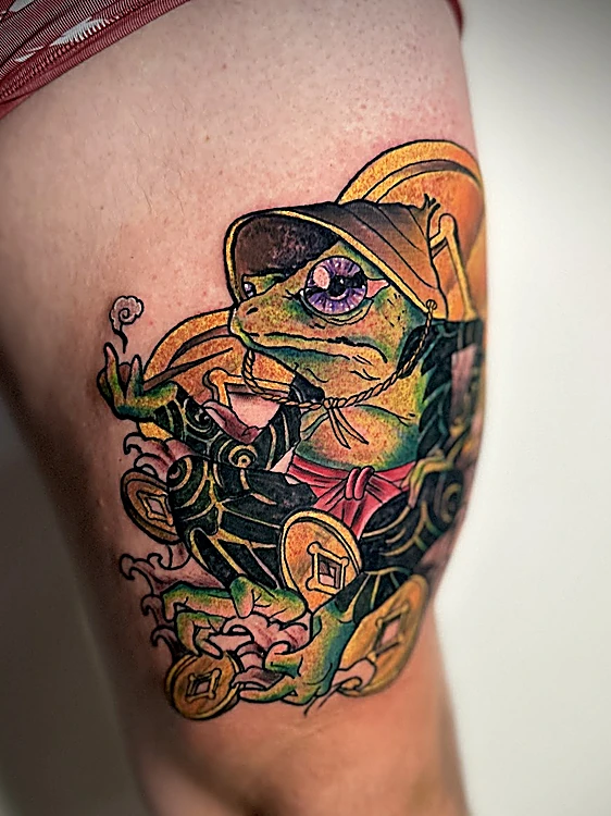 Tatuaje a color en el muslo: una rana con sombrero de paja rodeada de monedas doradas.