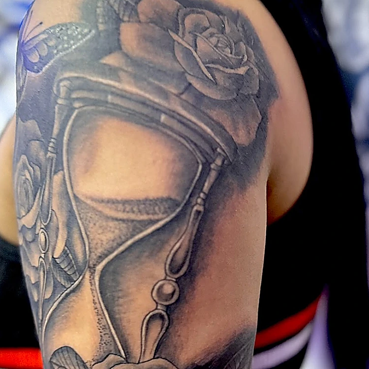 Tatuaje realista en negro y gris en el brazo: reloj de arena con rosas.