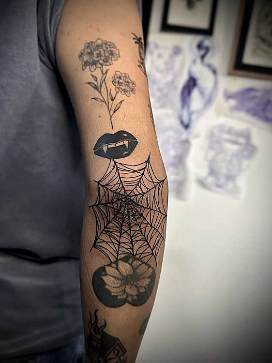 Brazo con varias piezas chicas: flores en línea fina, labios con colmillos y una telaraña en el codo; al fondo, hojas de flash.
